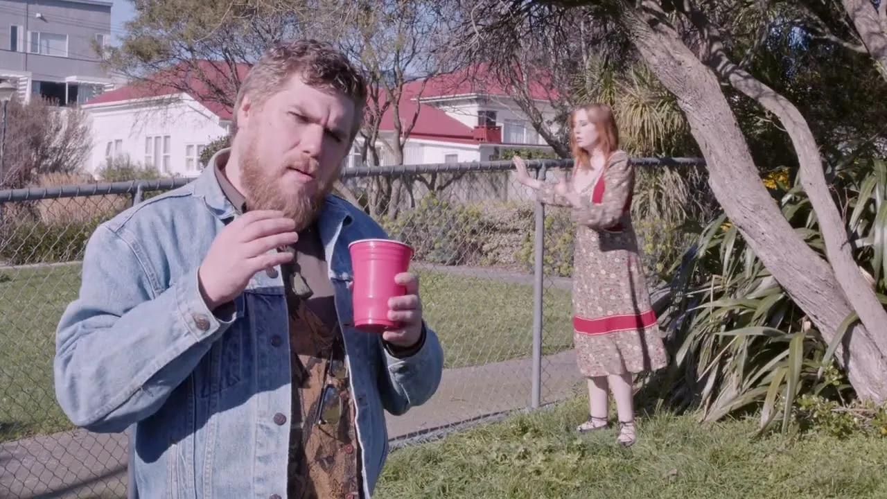 Still from Tongue Seekers: a man with a red cup by a fence while a woman watches